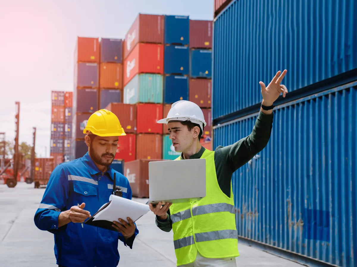 Zwei Experten in Warnweste und Helm prüfen mit Klemmbrett und Laptop die Abläufe im Logistikzentrum